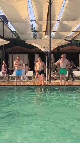 Three men in swim trunks jump into a swimming pool from a wooden deck. They are jumping one after another. The pool is surrounded by lounge chairs and cabanas. The scene is outdoors and appears to be during the daytime.