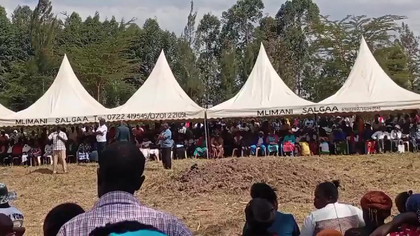 In a medium shot, a large group of people are gathered outdoors under three white tents. The tents, labeled "MLIMANI SALGAA," provide shade for rows of chairs where many people are seated. A few individuals stand in front of the seated crowd, seemingly addressing them. A man in a tan shirt appears to be speaking into a microphone. The camera is positioned behind a smaller crowd, giving a view of the event from a more distant perspective. The background features tall green trees, and the ground is uneven with patches of dirt. The overall atmosphere suggests an outdoor gathering or event, possibly a community meeting or celebration.