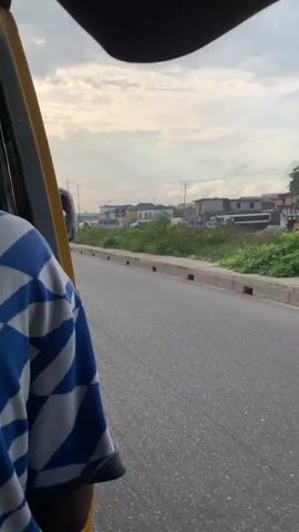 The video captures a view from inside a moving vehicle, likely a tricycle (keke), traveling along a paved road in Satellite Town, Nigeria. The top of the vehicle's frame, in yellow and black, is visible in the upper left corner. A person wearing a blue and white patterned shirt is partially visible in the foreground, suggesting they are a passenger.

The road features a concrete drainage ditch running alongside it, separating it from an overgrown grassy area. Beyond the grass, various buildings, including some residential structures and potentially commercial establishments, can be seen in the distance. Streetlights and power lines with pylons are visible along the roadside, indicating urban infrastructure. The sky is overcast with a light cloud cover, suggesting it is daytime but not sunny. No specific text is clearly discernible on any signs or buildings. The scene depicts typical roadside scenery in a developing urban area.