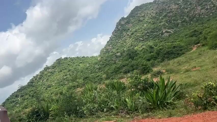 A tilted shot shows a hilly, green landscape under a partly cloudy sky. In the foreground, the right side of a yellow and green auto-rickshaw is visible, parked on a dirt road. The auto-rickshaw has a license plate that reads "PTA 6016". The background is dominated by a large, vegetated hill with scattered rocks. The camera appears to be moving slightly.
