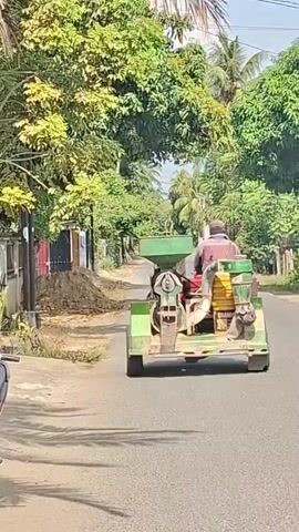 A person drives a green tractor with a milling machine attached down a paved road. The tractor has a large green hopper at the front and a yellow container at the back. The person operating the tractor is wearing a brown shirt and a hat. The road is lined with trees and houses. There is a pile of dirt on the side of the road. The sky is visible and appears to be clear. The overall environment suggests a rural or agricultural setting.