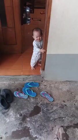 A toddler stands in a doorway, leaning against the doorframe. The toddler is wearing a white onesie with a gray pattern. Several pairs of flip-flops are scattered on the ground outside the doorway, along with a pair of black slide sandals. The floor outside appears to be concrete and is wet in several spots. The doorway leads into a room with tiled flooring.