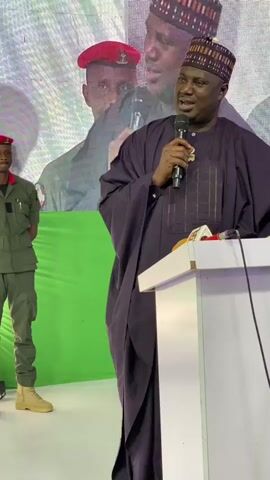 The video shows a man in traditional Nigerian attire, a dark purple robe and a patterned cap, speaking into a microphone at a podium. He is smiling and appears engaged with his audience. To his left, a man in a green uniform with a red beret stands at attention. Behind them, a large screen displays images of other men, some in uniform. A green backdrop is visible behind the man speaking and the uniformed man. Two microphones are on the podium, one of which the man is holding. The scene suggests a formal event or presentation taking place indoors, possibly a conference or political gathering. The lighting is bright, indicating it is likely daytime. No specific weather conditions or time of day beyond "daytime" can be inferred. The location is specified as Unguwar Rimi, Nigeria. The man speaking seems to be in a position of authority or is a prominent speaker at the event. His expression conveys a positive and approachable demeanor.
