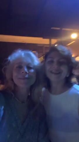 Two women are in a dimly lit outdoor venue. The woman on the left has blonde, wavy hair and is wearing a light-colored, possibly sequined top. She is smiling and looking at the camera. The woman on the right has dark brown hair and is wearing a white top. She is also smiling and looking at the camera. The background is blurry but appears to show foliage and lights, suggesting an evening outdoor setting. The lighting in the video shifts between purple and green hues, indicating the presence of colored lights. The overall atmosphere seems festive and celebratory.