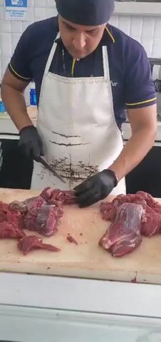 A man wearing a dark blue shirt, a white apron, and black gloves is cutting meat with a knife on a white cutting board. There are piles of raw meat on the cutting board. The man appears to be a butcher in a kitchen or food preparation area. The background shows stainless steel surfaces and what appears to be a tiled wall.