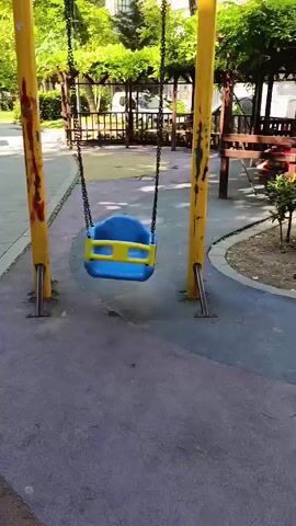 A swing set with a blue and yellow seat hangs stationary in a park setting. The swing set has yellow poles and a yellow horizontal bar. Trees and buildings are visible in the background. The ground is paved with purple and pinkish material. The lighting suggests it is daytime.