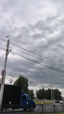 The video shows a street view with a dark, cloudy sky. A blue truck with a black cargo box is parked on the side of the road, partially obscured by trees and a fence. Overhead wires are strung across the frame. The scene appears to be in a suburban or semi-urban area, with some buildings and other vehicles visible in the background. The overall atmosphere is overcast and potentially windy, given the texture of the clouds.