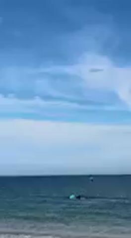 The video captures an expansive coastal scene featuring a person engaged in the sport of wing foiling in Barbate, Spain.

**Subjects:** The primary subject is a person actively wing foiling.
**Objects:** Key objects include a hydrofoil board, a handheld inflatable wing used for propulsion, the vast blue ocean, and a clear sky.
**Scene:** The scene is set on the open water of the ocean, stretching towards the horizon, with a small portion of a sandy beach visible at the bottom edge of the frame. The sky occupies a significant portion of the view.
**Setting:** The setting is a bright, sunny coastal area, confirmed as Barbate, Spain, which is known for its favorable conditions for water sports.

**Activity or event taking place:** The main activity is wing foiling. The person is skillfully riding a hydrofoil board across the water, propelled by a large, handheld inflatable wing. Throughout the video, they perform various maneuvers, gliding smoothly over the water, and occasionally lifting the board entirely out of the water due to the hydrofoil's lift and the wind's power.

**Notable details:**
*   **Time of day:** The video appears to be shot during the daytime, likely late morning or early afternoon, given the bright and consistent natural lighting.
*   **Weather:** The weather conditions are excellent for water sports, characterized by a clear, vibrant blue sky with scattered, fluffy white clouds. The ocean surface is relatively calm, with gentle ripples and small waves, indicating moderate winds necessary for wing foiling.
*   **Location cues:** The wide, open body of water and the distant coastline strongly suggest a coastal beach environment. The provided location context identifies it specifically as Barbate, Spain.
*   **Emotions:** The scene conveys a sense of adventure, skill, and enjoyment, reflecting the exhilaration of participating in a dynamic water sport in a beautiful natural setting.
*   **Visible text:** No visible text appears in the video.
