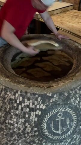 In an indoor setting, likely a bakery or restaurant in Tsovagyugh, Armenia, a person is seen baking bread in a tandoor oven. The tandoor, a large cylindrical clay oven, is decorated with dark and light mosaic tiles and features a circular emblem with an anchor and fork design on its exterior.

The individual, wearing a red shirt and a white cap, leans deep into the hot tandoor opening to skillfully press pieces of raw, flattened bread dough onto the interior walls of the oven. The dough adheres to the hot surface, beginning to cook and puff up. The camera captures the baker's precise movements and then provides a closer view of the numerous dough pieces arranged inside the oven.