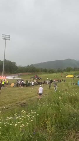 The video shows a group of people gathered in an outdoor grassy area with hills and trees in the background under a cloudy sky. There are several tents set up, possibly for an event, and some vehicles, including a yellow van. Some people are walking around, and others appear to be standing in groups. In the foreground, there are tall grasses and flowers. The overall atmosphere seems to be that of an outdoor gathering or event, possibly a race or festival, with a slight haze or overcast weather.