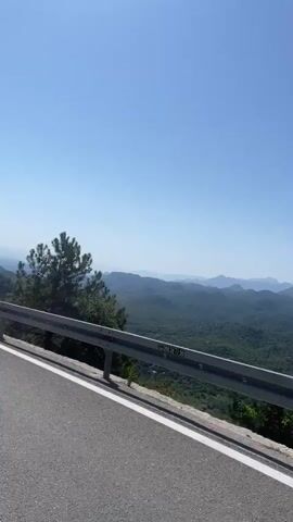 A POV shot from a vehicle driving along a mountain road shows a vast expanse of green, rolling hills and mountains under a clear blue sky. A metal guardrail runs along the edge of the road in the foreground, separating the road from the steep drop-off and the scenic vista. Trees, including pine trees and deciduous trees with green and orange foliage, are visible along the roadside and dot the hillsides. In the distance, the landscape fades into a hazy, lighter blue, suggesting a far-off body of water or the atmosphere. The video appears to capture a journey through a mountainous, possibly coastal, region on a sunny day.