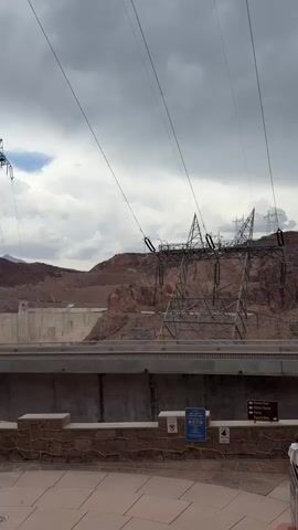 The video shows a view of a large dam structure with mountainous terrain in the background and a cloudy sky overhead. Several large power transmission towers are visible, some of which appear to be leaning or damaged. The foreground shows a paved walkway and some stone walls. A few people are walking on the walkway and stairs. There are also signs and trash receptacles visible. The overall impression is of a scenic location with some industrial infrastructure.