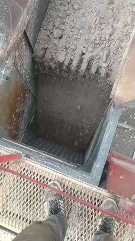 A first-person perspective video shows a person standing on a metal platform above a large industrial machine. The machine appears to be a crusher or grinder, filled with a large amount of grey, rocky material. The material is being processed, with smaller pieces falling into the machine. The person's feet and legs are visible in the foreground, wearing dark pants and athletic shoes. The environment appears to be an industrial setting, likely a construction site or quarry. The lighting is bright, suggesting daylight conditions. The camera is pointed downwards into the machine.
