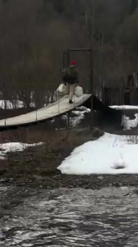 A video shows a river flowing with snowy banks. A person with a red hat, wearing a jacket and light pants, walks across a small, partially covered bridge, carrying two white bags. The trees are bare and the setting appears to be rural.