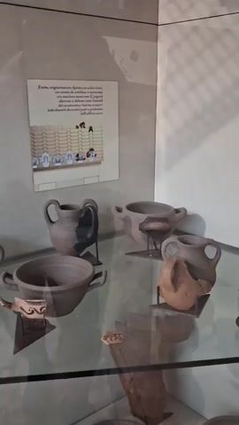 The video shows a museum display case with various ancient artifacts. The display case has a glass top and is filled with pottery fragments, tools, and what appear to be weights or spools. On the wall behind the display case are two informational plaques with text and images, one depicting what looks like a manuscript or illustration and the other showing a pair of bronze scissors. The lighting in the museum is dim, with shadows cast on the wall. The camera is positioned to capture a close-up view of the artifacts within the display case.