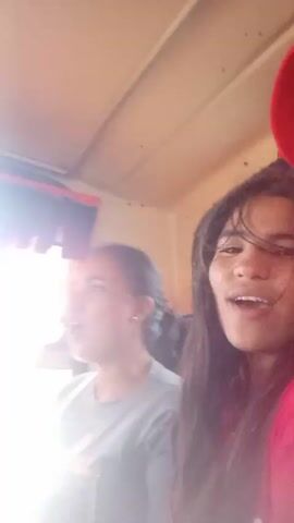 Two young women are on a boat, singing along to loud music. The woman on the left is wearing a grey t-shirt, and the one on the right is in a red shirt. They both look like they are enjoying themselves. The boat interior is visible, with a window and curtains.