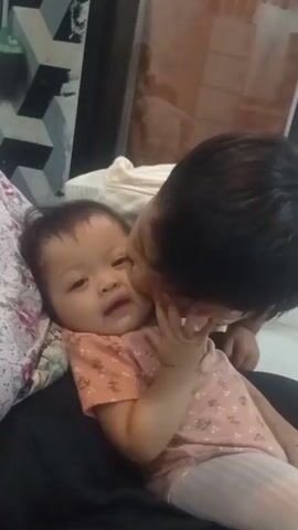 In the foreground, a baby is lying down with its head slightly turned to the left, looking at the camera with a slight smile. The baby is wearing a short-sleeved pink shirt with a floral pattern and light-colored pants. A person, possibly an older sibling, is leaning in from the right and kissing the baby on the cheek. The older sibling has dark hair and is wearing a light-colored t-shirt. The background is blurred, but appears to be an indoor setting with a wall featuring a geometric pattern and a large mirror or glass surface. The scene is brightly lit.