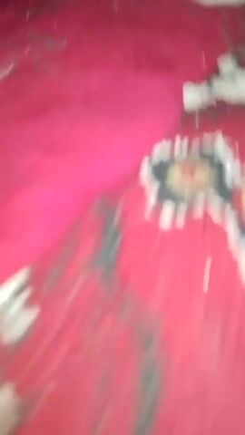 A white and gray cat wearing a red collar is lying on a red surface. The cat's eyes are closed and it appears to be sleeping. The surface has a textured appearance, with some small white flecks scattered across it. The lighting is dim, and the camera is positioned close to the cat. The image is slightly blurry.