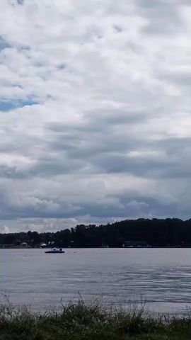 A wide shot of a large body of water is captured under a cloudy sky. The water is calm, reflecting the gray and white clouds above. In the distance, a tree-lined shore is visible, dotted with houses and other structures. A small boat is moving across the water towards the shore. In the foreground, tall grass and foliage frame the bottom of the shot. The overall atmosphere is peaceful and serene, with a sense of open space and natural beauty.