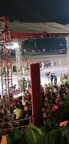 The video shows an outdoor night event with a stage set up. In the foreground, there are spectators seated and some people standing behind them. A large metal truss structure, likely part of the stage lighting or sound system, is prominently visible on the left. Further back, on an open paved area, a large group of people in uniform are assembled in formation. Some individuals in military-style uniforms are standing on a raised platform or stage area in the middle ground, interacting with each other. A camera on a tripod is also visible in this area, suggesting a recording is taking place. Behind the uniformed individuals, another large group of people in red and green uniforms are standing in formation. The overall impression is of a formal ceremony or parade.