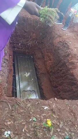 A priest in a purple robe stands over a grave with a coffin inside and throws a bundle of leafy plants into the grave. Then, someone in white holds a blue basin of water, and water is poured into the grave. Other people surround the grave. The ground is dirt and has a few flowers nearby.