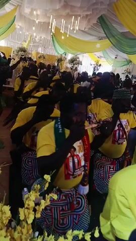 A group of dark-skinned men wearing yellow shirts and patterned wraps play reed instruments in unison. Most of the men have a laminated badge hanging from their necks and are holding the instruments horizontally to their mouths. The scene takes place indoors beneath a white, yellow, and green draped ceiling. There are other people in the background watching.