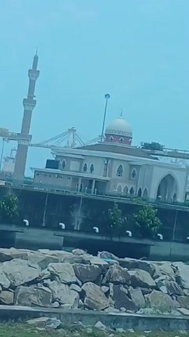 A panning video shows a waterfront with a mosque and minaret. In the background, there are shipping cranes and a cargo ship. The foreground consists of a rocky shoreline and a grassy area. The sky is clear and blue.