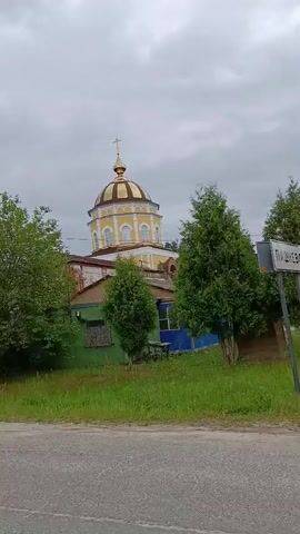 The video captures a view of a church complex in Gribchikha, Russia. The scene is dominated by a tall, yellow and white bell tower with arched openings, topped with a golden dome and cross. To the right of the tower, a smaller building with a golden dome, also adorned with a cross, is visible. In the foreground, lush green grass lines a paved road. Various trees and bushes surround the buildings, suggesting a rural or semi-rural setting. The sky is overcast and cloudy, indicating a likely gray or possibly rainy day. No people are visible in the footage, and the focus is solely on the architectural elements of the church and its natural surroundings. The overall mood is serene and tranquil, highlighting the architectural beauty and the peaceful atmosphere of the location. The time of day is not definitively ascertainable due to the cloudy conditions, but it appears to be daytime. No text is visible in the video.