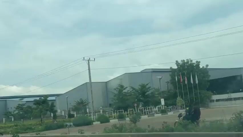 The media file depicts an outdoor scene in Kwali, Nigeria, under a cloudy sky, suggesting it is either early morning or late afternoon. The primary subject is a large, modern industrial building with a grey, corrugated metal exterior. The building appears to be a factory or warehouse, with several sections and a prominent roofline.

In the foreground, several people are engaged in various activities. One shot shows two individuals on a motorcycle driving past the building, hinting at transportation or movement within the area. Another shot focuses on a silver hatchback car with its trunk open. A man in a yellow shirt and a woman in a patterned dress are busy loading items into the trunk. A third woman, wearing a blue headscarf, sits on a concrete barrier nearby, holding a shopping bag with "SAS" visible on it. This suggests an activity related to shopping, delivery, or personal errands.

Notable details include utility poles and power lines running across the frame, common in many built-up areas. Trees and landscaping are visible around the industrial building, indicating some attention to the surrounding environment. A flagpole with several flags can be seen on the right side of the industrial building, but their designs are not clearly discernible. The overall mood appears to be one of everyday activity rather than a specific event. The overcast weather contributes to a subdued lighting condition.