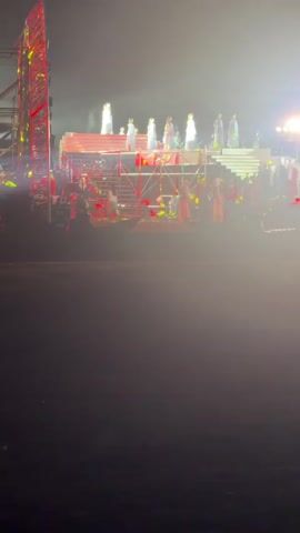 The video shows a performance stage at night. There are several people standing on a raised platform at the back of the stage, all wearing white. In front of them, there is a structure with red lights and stairs. Some people are visible on the stairs and around the structure. There are also lights shining on the stage from the sides, creating a hazy effect. The ground in front of the stage is dark, with some blurred green lights visible in the lower left corner. The overall atmosphere suggests an outdoor event or concert.
