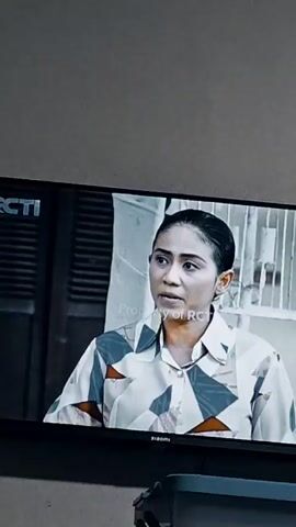 A woman with dark hair pulled back smiles and looks to her right. She is wearing a patterned collared shirt. Behind her, a shadowy background features architectural elements and what appears to be a birdcage. The text "PROPERTY OF RCTI" is overlaid on the image. The scene then changes to show a woman wearing a headscarf and a patterned jacket standing next to a man wearing a light-colored t-shirt and a dark cap. The man is holding a birdcage. They are indoors, in what looks like a living space. The woman is looking to her left with a concerned expression, while the man is looking up and to his right with his mouth open, as if speaking.