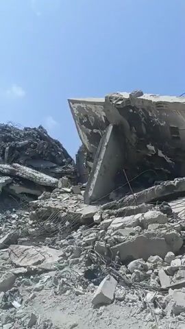 The video shows a massive pile of rubble, consisting of broken concrete slabs, twisted metal, and debris, under a bright blue sky with scattered clouds. The scale of the destruction suggests a building or structure has collapsed. The camera pans slowly across the scene, highlighting the jagged edges of the concrete and the exposed rebar. There are no people or any other objects clearly visible in the frame, nor are there any apparent actions or interactions taking place. The environment appears to be an outdoor, possibly urban, setting.
