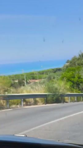 The video shows a view from inside a vehicle traveling on a paved road that curves to the right. The road has a white line marking the center. On the left side of the road, there is a metal guardrail, followed by lush green vegetation, including tall grass and trees. In the distance, beyond the vegetation, a bright blue body of water, likely the ocean, is visible under a clear blue sky. On the right side of the road, there are trees and shrubs, with some areas of rocky terrain visible. Two cars are seen on the road ahead. The first is a white car parked on the left shoulder of the road, partially obscured by trees. The second is a silver SUV, further ahead, traveling in the same direction as the viewer. The video captures a sunny day with clear visibility.
