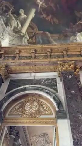 The video shows an upward view of an ornate ceiling painting within an elaborately decorated building, potentially a palace or museum. The painting is a classical-style fresco depicting numerous figures, including cherubs, deities, and human-like characters in a cloud-filled sky. The colors are predominantly soft pastels with bursts of red, blue, and gold. Below the painting is an ornate gold-trimmed border and a marble arched doorway with additional gold detailing. The camera slowly pans upwards, focusing on the details of the ceiling painting.