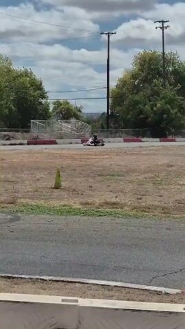 The video shows a go-kart race on a sunny day. Several go-karts with drivers wearing helmets are visible on a paved track with grassy areas and trees. An American flag flies on a flagpole in the foreground. The karts are moving at speed, and the video captures them from various angles as they navigate the track. The environment appears to be an outdoor racing facility.