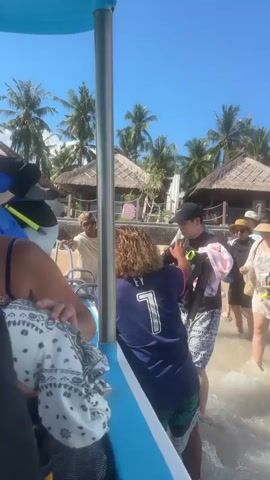 The video shows a group of people on a boat, with several others standing on the beach. The boat is small, open, and painted blue and white. People are boarding the boat from the sandy beach, which is lined with tropical palm trees and small thatched-roof huts. Some people carry snorkeling gear, including masks, fins, and snorkels, suggesting they are about to engage in water activities. The sky is clear and blue, indicating sunny weather. The people on the boat are dressed in casual beach attire, and those on the beach seem to be assisting with the boarding process.
