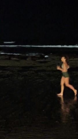 The video shows a woman jogging barefoot on a dark beach. She is wearing a sports bra and shorts, and her hair is pulled back. The water is calm with some visible waves in the background. The scene is dark, with limited light reflecting off the wet sand.