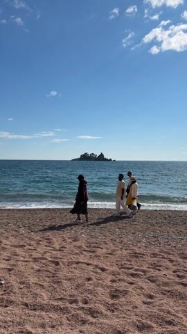 Four people are walking along a beach. The beach has reddish-brown sand or pebbles. The sea is blue, with small waves washing onto the shore.  A small island is visible in the distance. The sky is blue with a few scattered clouds.  The person on the left wears a dark patterned dress. The other three people wear light-colored clothing, including white and yellow.  The people are walking along the edge of the water.