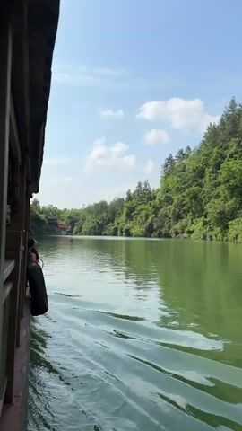 The video depicts a view from a moving boat on a river. The water is green, and the shoreline is covered in dense green trees and vegetation. The sky is blue with scattered white clouds. The boat's structure is visible on the left side of the frame, featuring dark wood and a black tire hanging on the side. In the distance, a red structure, possibly a bridge or building, can be seen. The camera is static, but the movement of the boat creates a sense of motion.