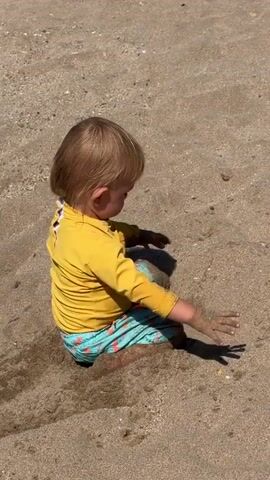 The video shows a toddler playing in the sand on a beach. The child, with blonde hair, is wearing a yellow long-sleeved rash guard and turquoise patterned swim trunks. They are kneeling and digging in the sand, with their hands moving in and out of the sand, creating shadows. The sun appears to be high in the sky, indicating a bright, sunny day. The sand is light brown with small pebbles and shells visible. The child's focus is on their activity, suggesting concentration and enjoyment. The scene is set at Sérignan, France, a location known for its beaches. No visible text is present in the video.
