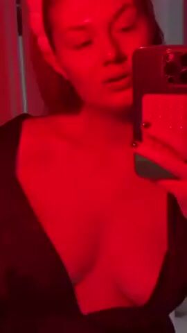 A woman with reddish hair is taking a selfie video in a room bathed in red light. She is wearing a black top that is unbuttoned to reveal her chest. She has a light-colored headband on. She holds a dark-colored phone with a textured design on the back. She points a finger and then puts her finger in her mouth. Music is playing in the background. There are some vertical lines visible in the background, possibly part of a door or wall.