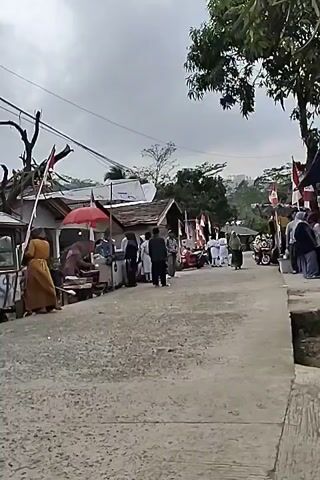 The video shows a street scene with several people and stalls. The weather appears overcast. Several red and white flags, possibly national flags, are displayed on poles along the street. There are temporary structures that appear to be market stalls, with awnings and goods displayed. People are seen walking and interacting near the stalls. A scooter is visible in the foreground, with a rider on it. Another person is walking away from the scooter. The overall impression is that of a local market or community event.