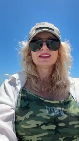 This is a selfie video taken from a woman's point of view at a beach. The woman is wearing a camouflage baseball cap with an American flag patch, sunglasses, a white open button-down shirt over a camouflage swimsuit with the word "PINK" embellished on it. Her blonde hair is visible around her face, and she is smiling. The background shows a bright blue sky and a turquoise ocean with waves. Several people are visible in the water in the distance, some swimming and others standing. The camera is at chest level and appears to be slightly submerged in the water, as indicated by the ripples and the angle of the shot. The overall scene suggests a sunny day at the beach.