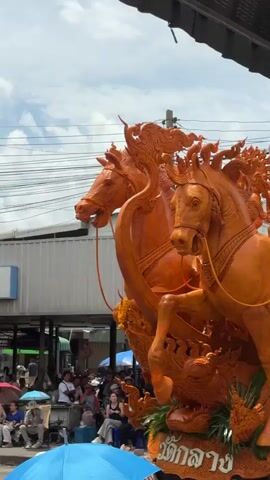 The video shows a close-up of a large, ornate orange sculpture of two horses in profile, one in front of the other, with intricate details and embellishments. The horses appear to be made of wax, likely part of a parade float or religious display. The sculpture is positioned outdoors, with a hazy sky and overhead wires visible in the background. In the lower portion of the frame, glimpses of people are visible, some seated, holding umbrellas, and others standing, suggesting a public event or festival. The overall scene has a festive and cultural atmosphere.