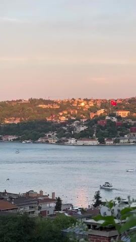 This is a vertical, high-angle shot overlooking a body of water, likely the Bosphorus Strait in Beşiktaş, Turkey. The time of day is sunset, as indicated by the warm, soft light illuminating the sky with hues of pink, orange, and pale yellow. The water reflects this warm light, creating a shimmering surface.

The scene features a hilly coastline dotted with houses and buildings, some with red-tiled roofs, nestled amongst lush green trees. The architecture suggests a Mediterranean or Eastern European influence. A prominent Turkish flag, red with a white star and crescent, flies from a flagpole on the hillside. Several boats are visible on the water, including a ferry or large passenger boat closer to the foreground and smaller vessels further out. A single bird is captured in flight over the water.

The overall mood of the scene is peaceful and picturesque. The soft lighting and the serene water evoke a sense of tranquility and beauty. The presence of the Turkish flag strongly suggests the location is Turkey. The image captures a typical sunset over the Bosphorus, a renowned landmark. There is no visible text in the media.