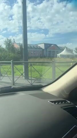 The video is shot from the interior of a vehicle, looking out through the windshield. The vehicle appears to be moving along a road bordered by green grass and trees. In the background, there is a large building with a white facade and a grey roof, possibly a mansion or a government building. There are also tents visible, suggesting an outdoor event. The sky is blue with fluffy white clouds. The video briefly shows a metal fence with a diamond pattern and a pedestrian crossing. A bus stop with people sitting on a bench is also visible. The sun is shining, creating a bright atmosphere and some glare on the windshield.