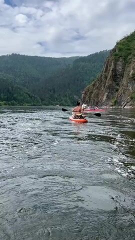 A person with a dog on a paddleboard is shown on a river. The person is sitting and paddling. There are other people on paddleboards in the background. The river is surrounded by trees and cliffs. The sky is cloudy.