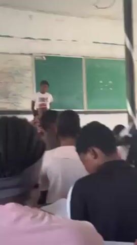 The video shows a classroom setting with students seated and a woman standing in front of the class. Behind her are two chalkboards, one covered with writing and the other with some equations. A man in a traditional patterned outfit and a cap sits in the front row, watching the woman. The camera is positioned behind the students, offering a view of the back of their heads. The classroom appears to be in a school.