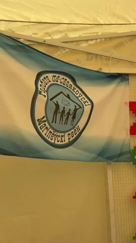 The video shows a close-up of a flag with a logo. The flag has a white background with a blue and white gradient design at the top and bottom. The logo in the center is circular and appears to be a crest or emblem. Inside the circle, there are silhouettes of people holding up a roof-like structure. There is text written around the circle in a dark color, which is difficult to read due to the resolution and angle. The flag is hanging, possibly from a tent or canopy, as indicated by the yellow fabric and metallic supports visible at the top. Part of a white and red striped object is visible on the right side, and a yellow surface is below the flag. The lighting is bright, suggesting it is daytime.