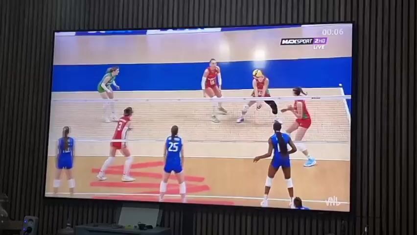A large flat-screen television, mounted on a wall with vertical wooden slats, displays an indoor women's volleyball match. The screen occupies the majority of the frame.

On the screen, two teams are competing. One team wears red jerseys with white numbers (e.g., number 19, 3, 18), and the other wears blue jerseys with white numbers (e.g., number 6, 25, 3, 5, 4). There are also players or officials visible in green/yellow attire. The players are in motion, with several positioned around the net, some jumping to block or hit the volleyball. The court features a light-colored playing surface, blue out-of-bounds areas, and distinct white boundary lines. Red arrow-like markings are also visible on the court floor. A volleyball net stretches across the middle of the court, and a referee's stand is visible to the right of the net.

Visible text overlays on the screen include "1XBET" repeated across the top, "MAXSPORT 2HD LIVE" with a timer "00:06" in the top right corner, "PIRACANJUBA PROFORCE" on the bottom left, and a "VNL" logo on the bottom right.

Below the television, partially visible in the foreground, are various electronic devices on a surface, including what appear to be speakers and possibly remote controls. The setting is an indoor space, likely a living room, with the wall texture providing a decorative element. The activity is a live or recorded professional volleyball match, captured with a high-angle camera view of the court. The players exhibit focus and competitive intent.